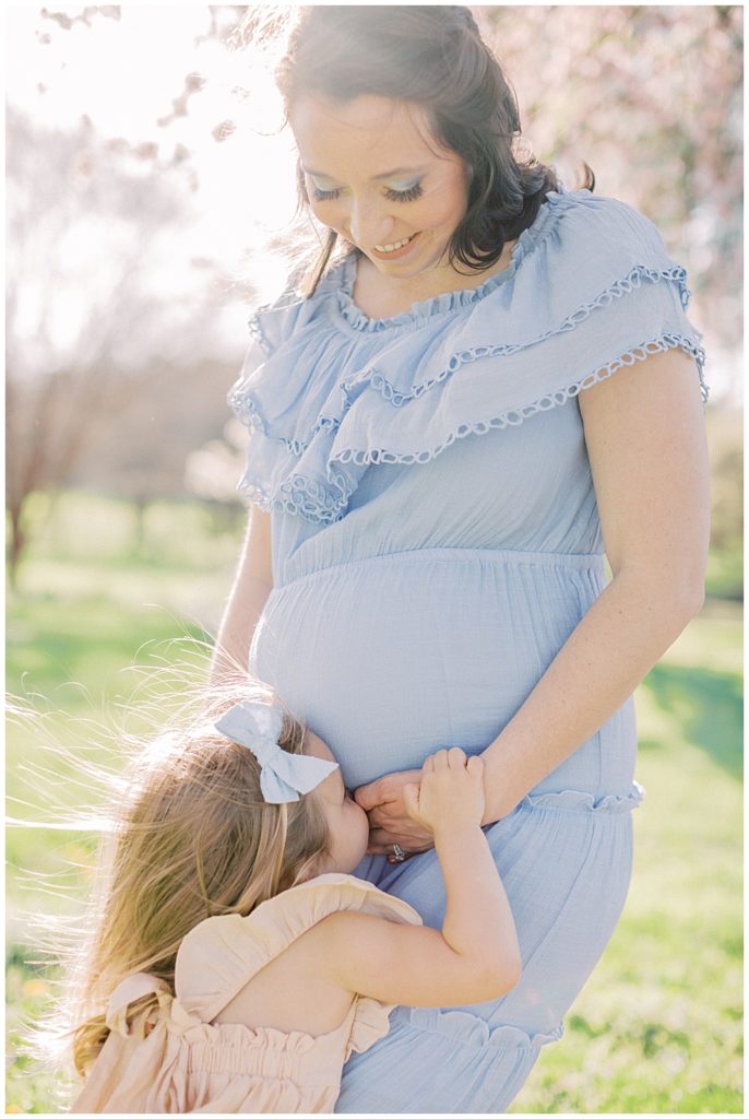 A toddler girl kisses her mom's pregnant belly during their maternity session at the National Arboretum