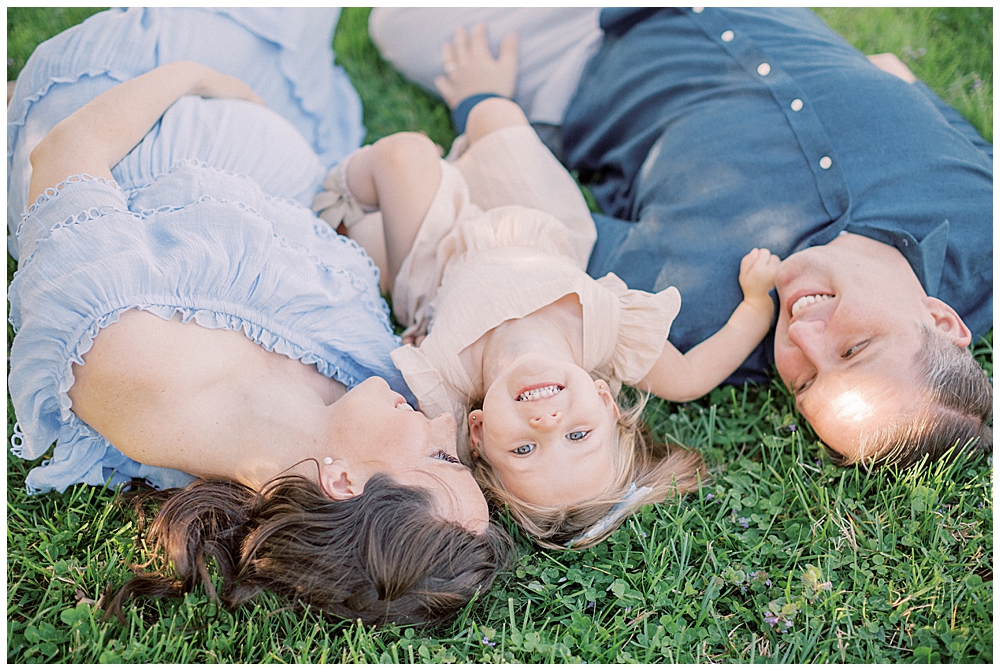 Mother, father, and toddler daughter lay down in the grass and smile