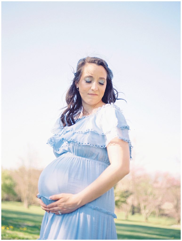 A pregnant mother wearing a blue dress stands with her eyes closed, holding her belly.