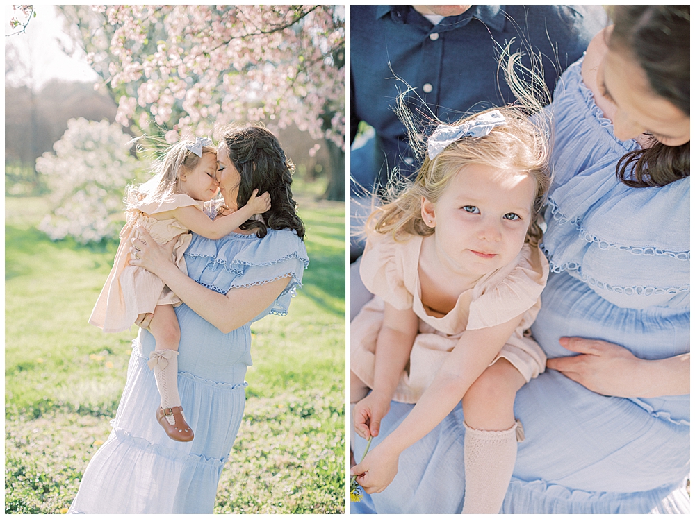 A little girl cuddles with her pregnant mother who is wearing a blue dress by a cherry tree