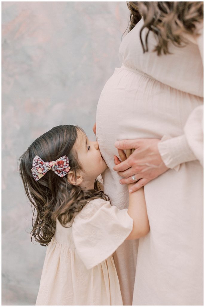 Little girl kisses her mother's pregnant belly during their maternity studio session with daughter and mother
