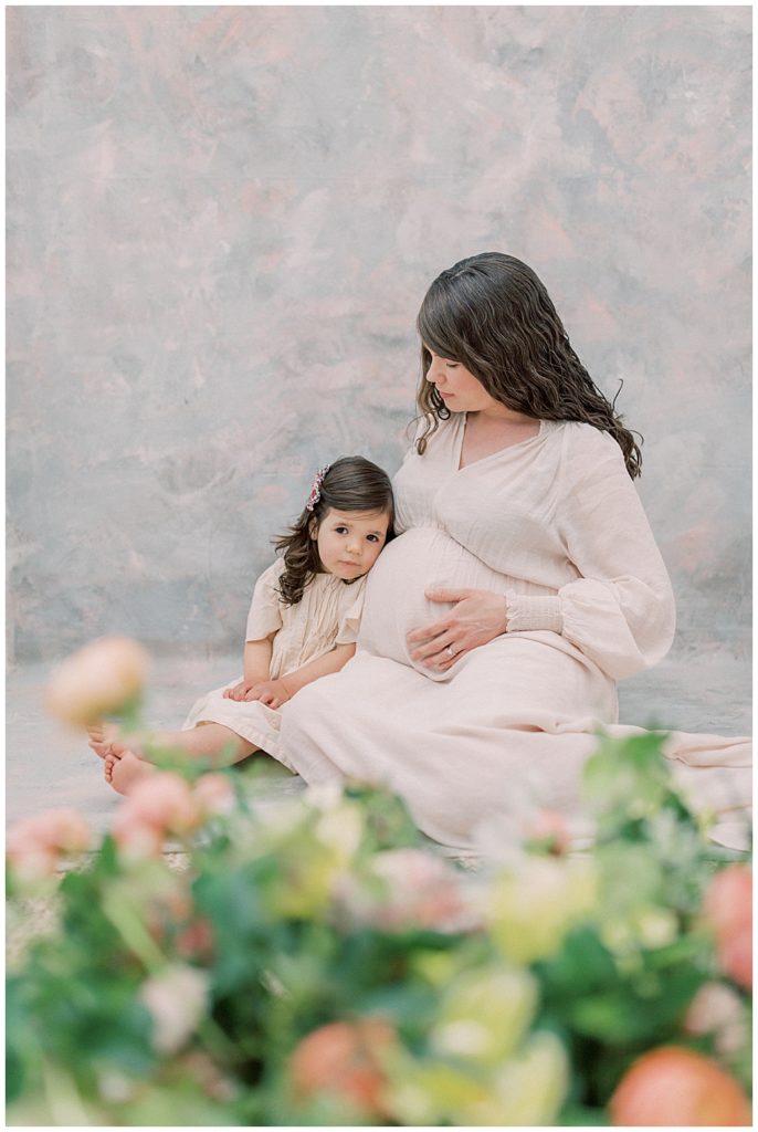 Pregnant mother and little girl sit in front of a floral display in a photography studio and the little girl lays her head on her mother.