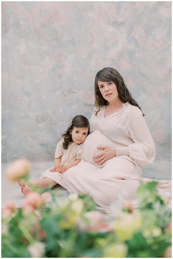 Mother and daughter sit in a photography studio in front of a floral display in the foreground and look at the camera during their Maternity Studio Session With Daughter and Mother