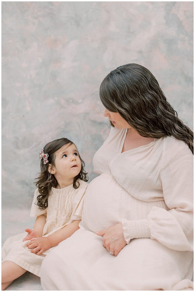 Toddler girl sits with her mother and looks up at her during Maternity Studio Session With Daughter and Mother