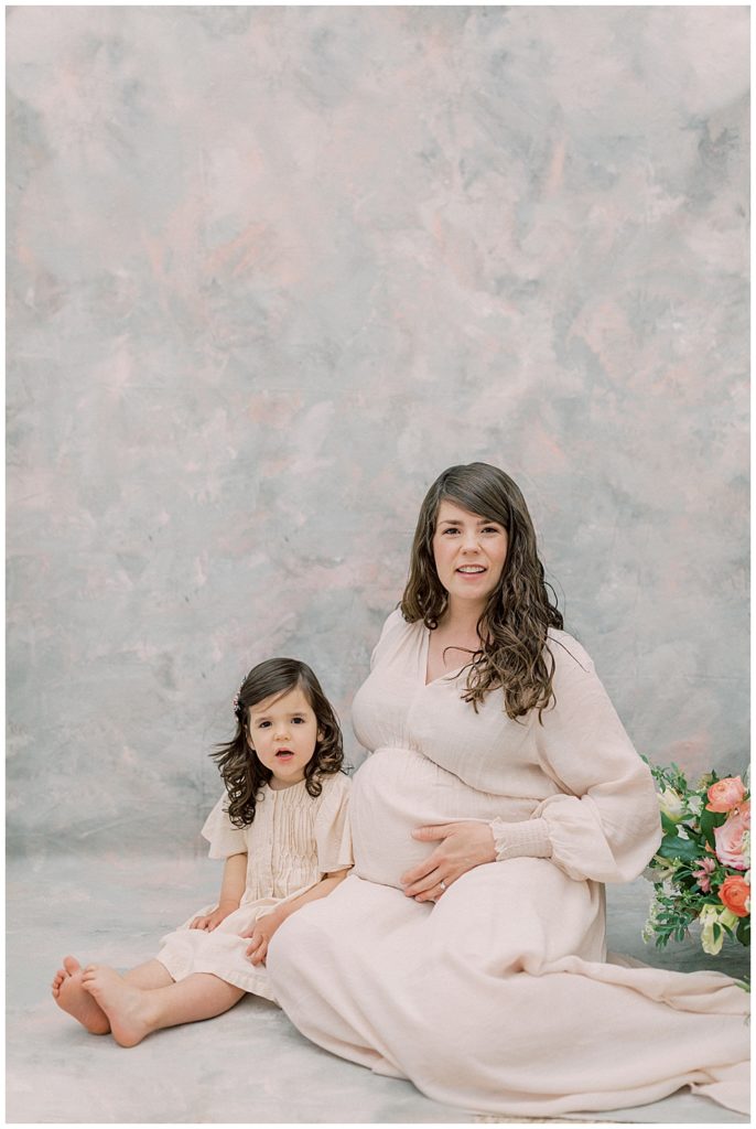 Pregnant mother sits with her little girl next to flowers in a photography studio during their maternity session