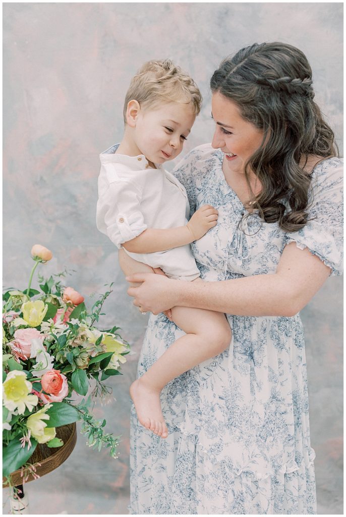 A mother wearing a blue Baltic Born dress holds her toddler son and smiles at him during their studio motherhood session