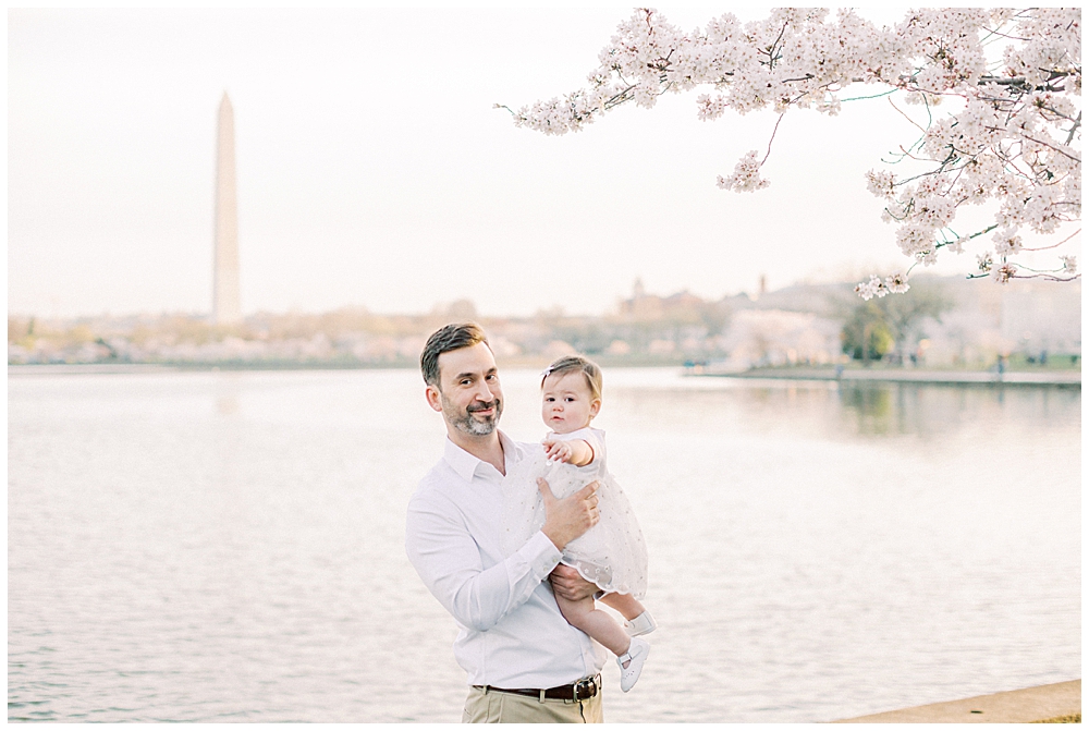 A father holds his baby daughter next to a cherry blossom tree across from the Washington monument in DC