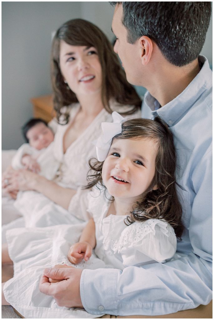 A new family of a mother, father, toddler daughter, and newborn daughter sit on a bed