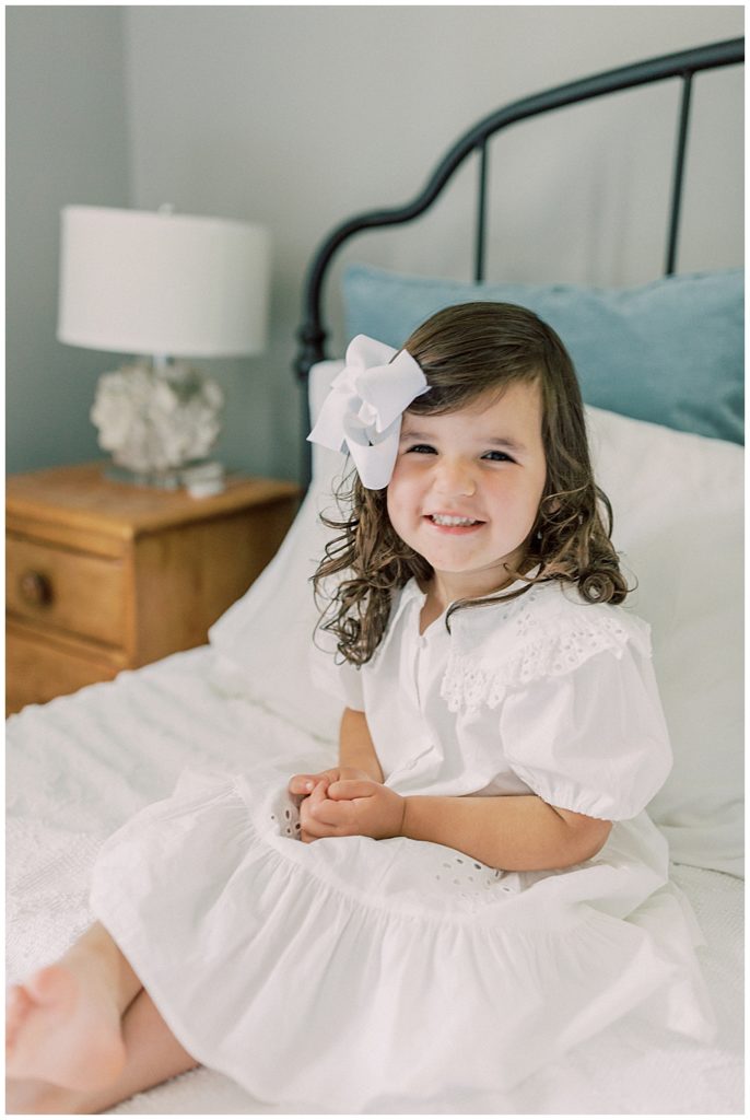 A toddler girl sits on the bed with her hands in her lap smiling