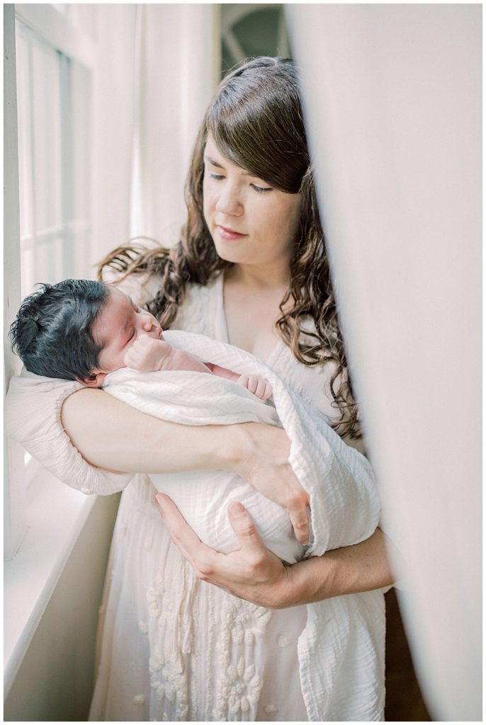 A mother holds her newborn daughter through a curtain by the window