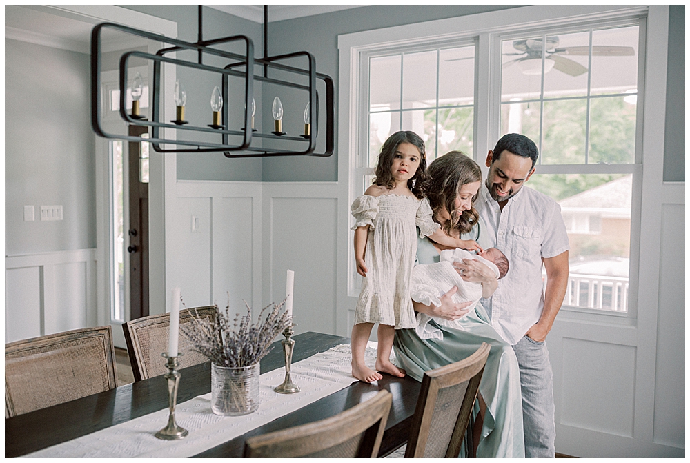 Mother, father, and toddler daughter sit on their dining room table while holding their newborn baby