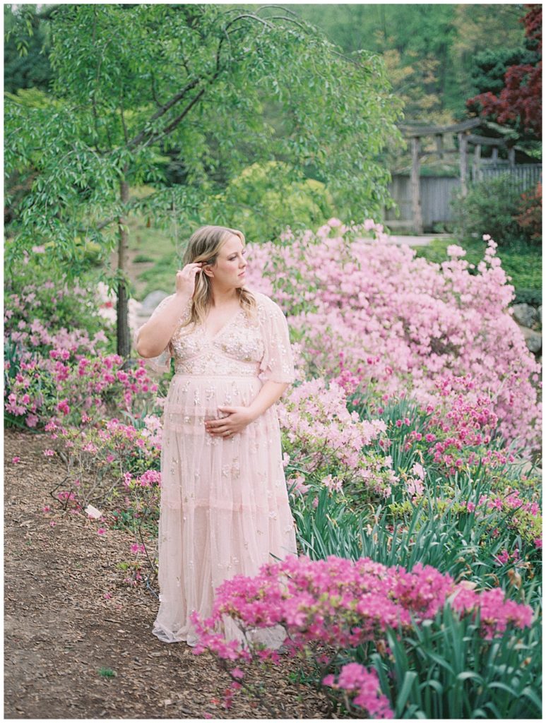 Pregnant woman with blonde hair and pink Needle & Thread gown stands in a garden with pink azaleas with one hand on her belly and one brushing her hair away.