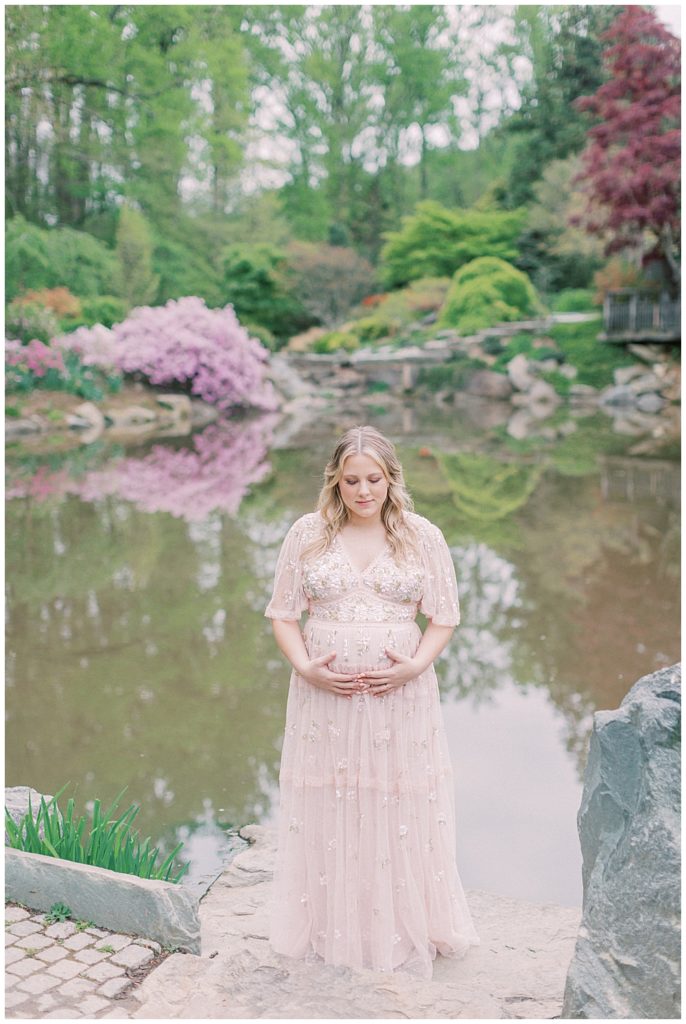 A blonde mother stands on a rock near the pond at Brookside Gardens while wearing a pink Needle & Thread gown.
