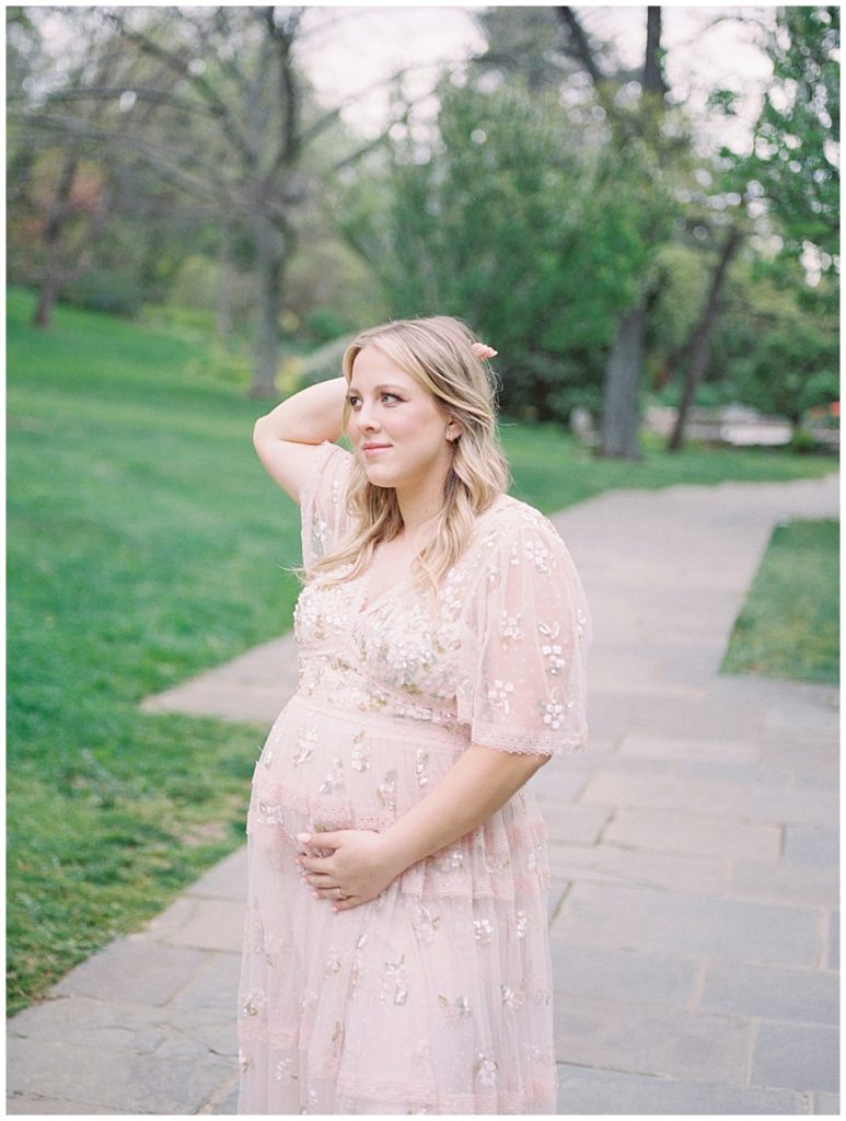 A mother with blonde hair has one hand on her belly and one behind her head during her garden maternity session at Brookside Gardens.