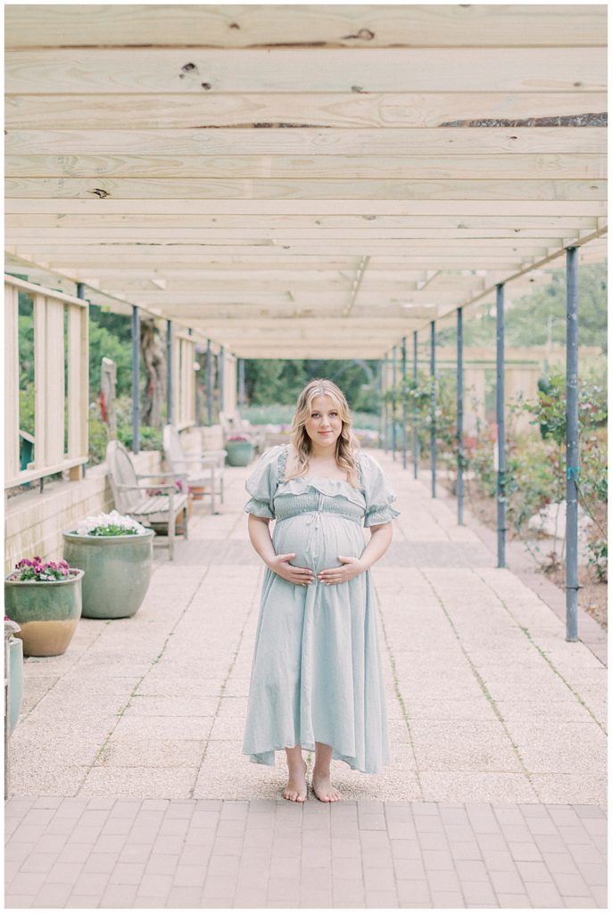 A pregnant blonde mother in a green dress stands with her hands below her belly while standing under a wooden pergola. 