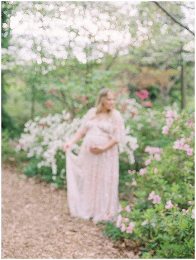An out-of-focus image of a pregnant mother in a pink dress holding her dress and cradling her belly in a garden.
