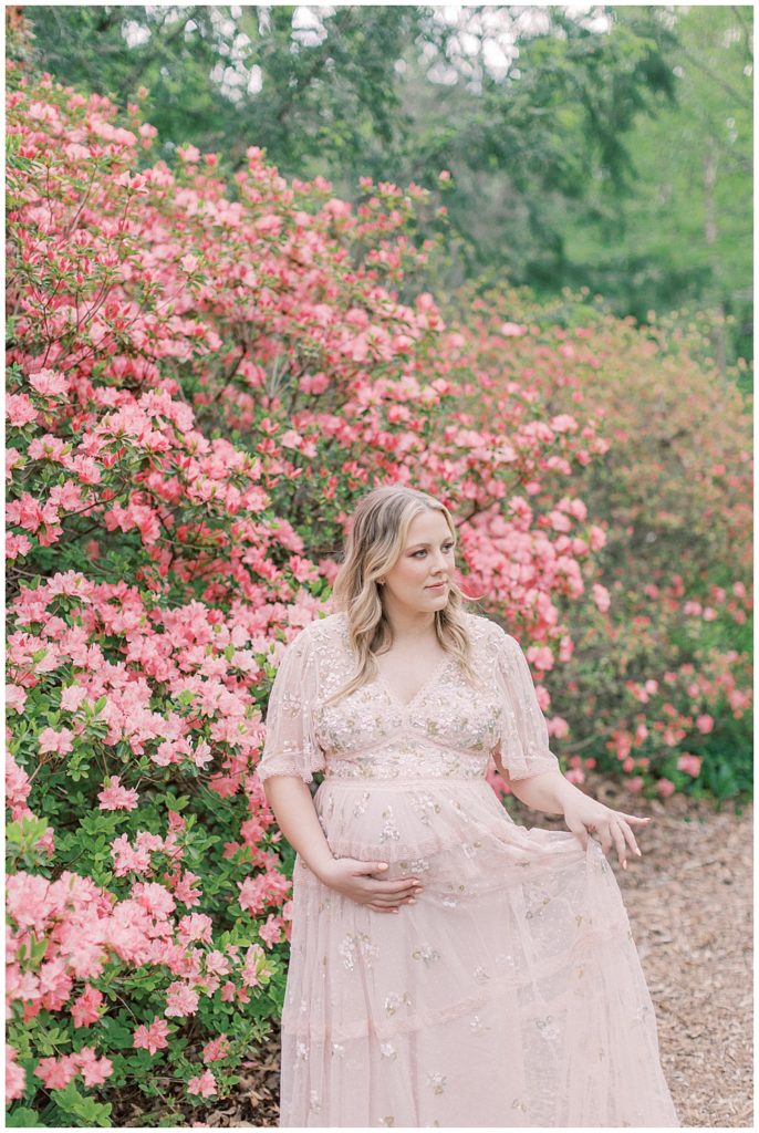 A blonde pregnant woman in a pink Needle & Thread dress stands near a pink azalea bush in Brookside Gardens during her maternity session