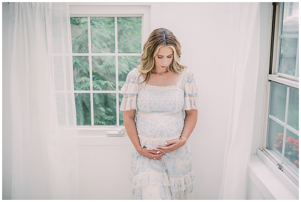 An expecting mother in a blue Needle & Thread dress cradles her pregnant belly and looks down during her studio maternity session.