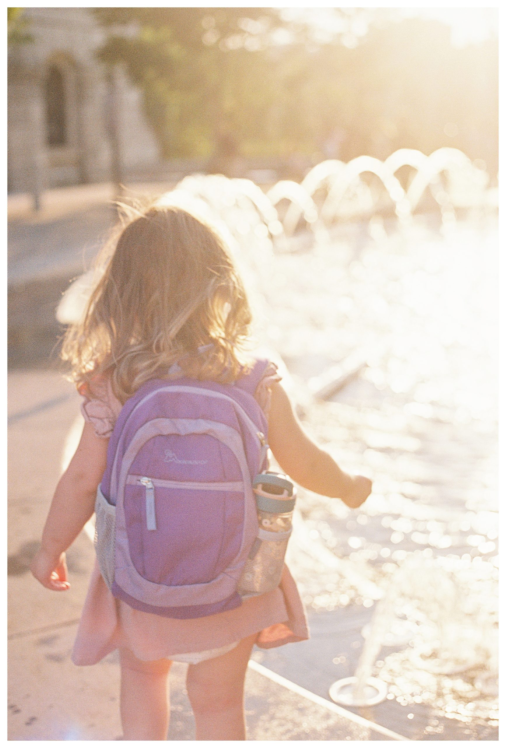 Little girl walks along fountain at sunset with purple backpack.