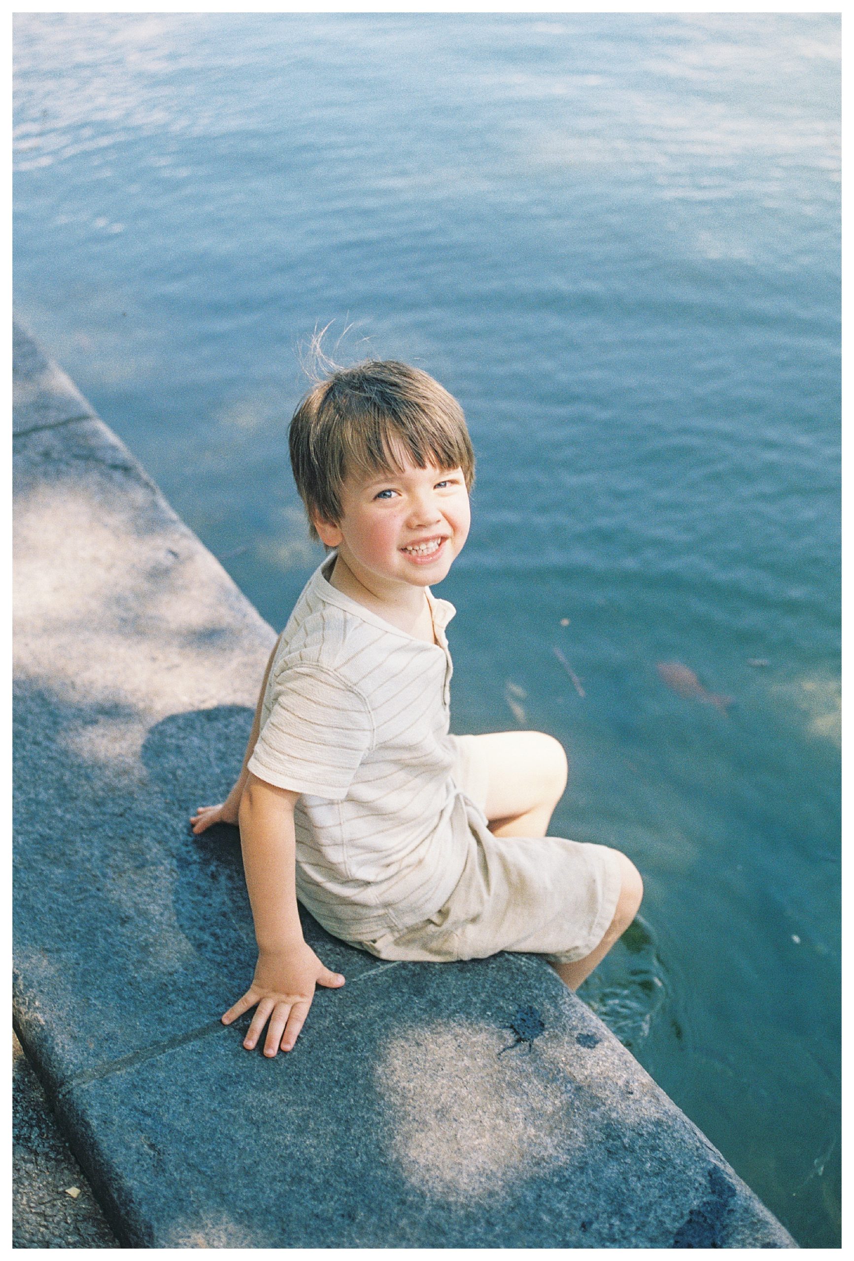Toddler boy with blonde hair sits on the edge of a lake looking up and smiling