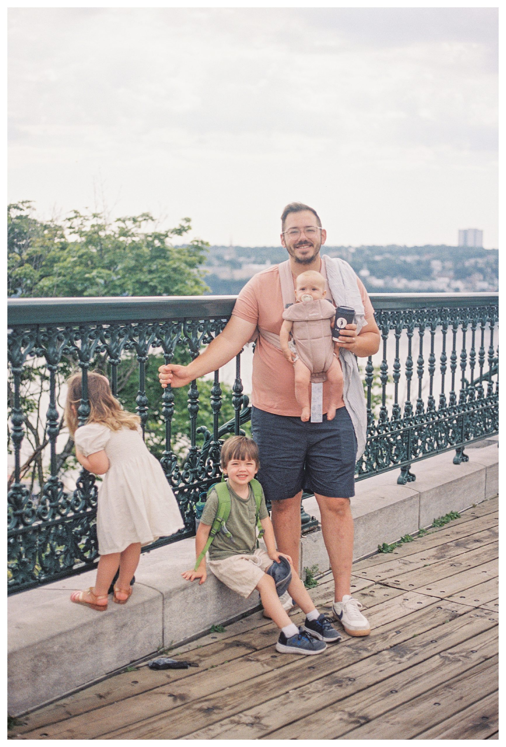 Father holding infant daughter in carrier stands with toddler son and toddler daughter as she looks through iron railing in Quebec City.
