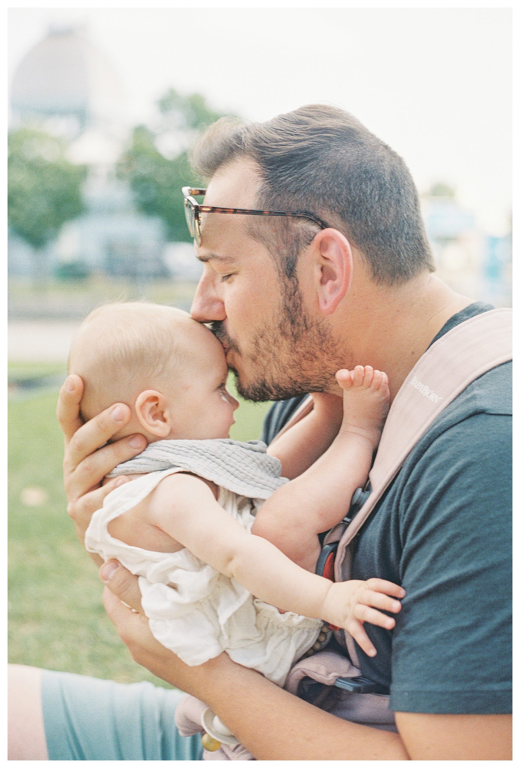 Father leans down to kiss his infant daughter on the forehead while sitting in a park.