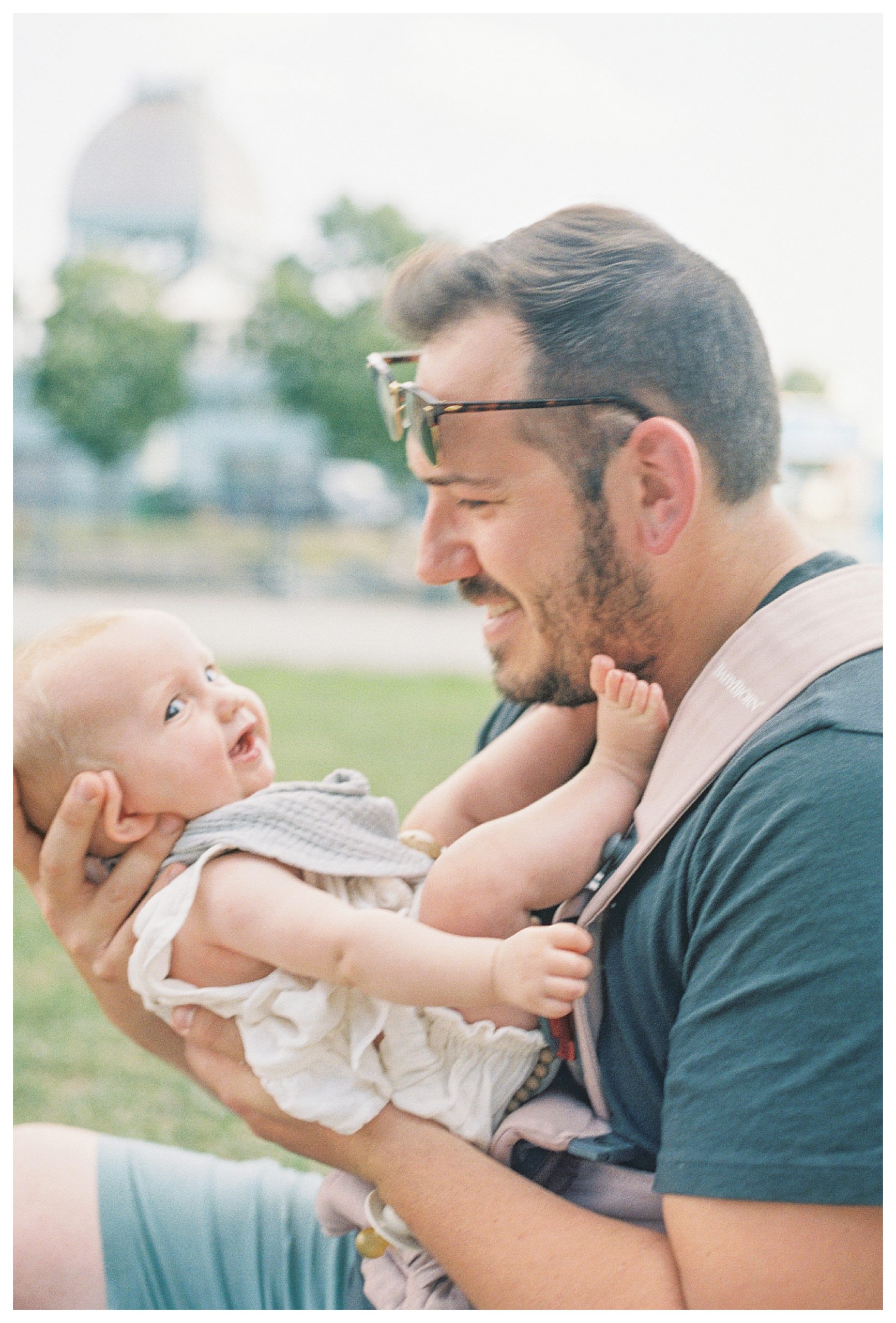 Father holds his infant daughter in front of him and smiles while sitting in a park.