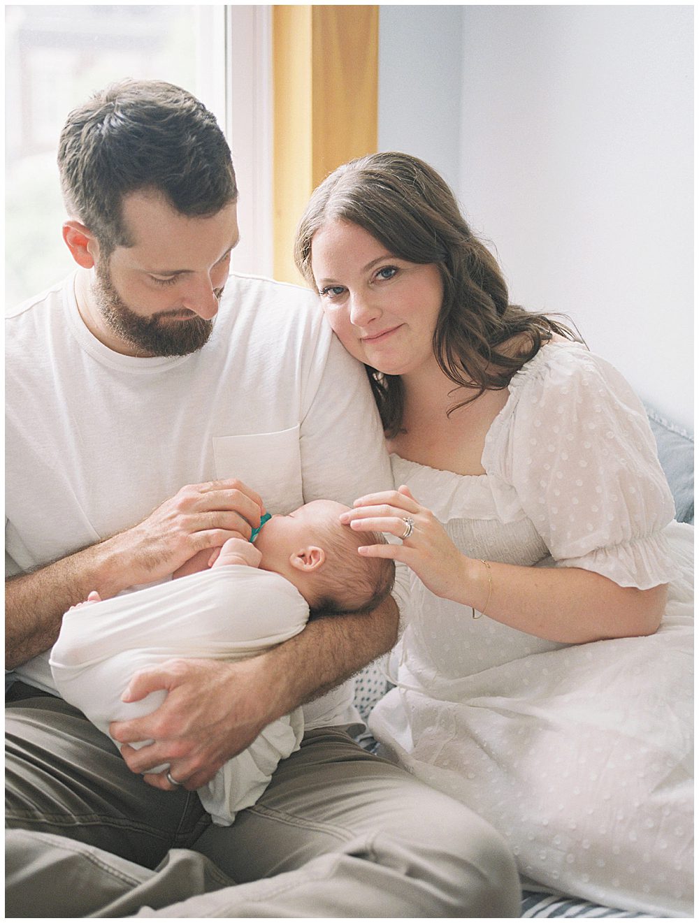 Mom looks at the camera with a soft smiles and touches her baby's head while her husband holds baby.