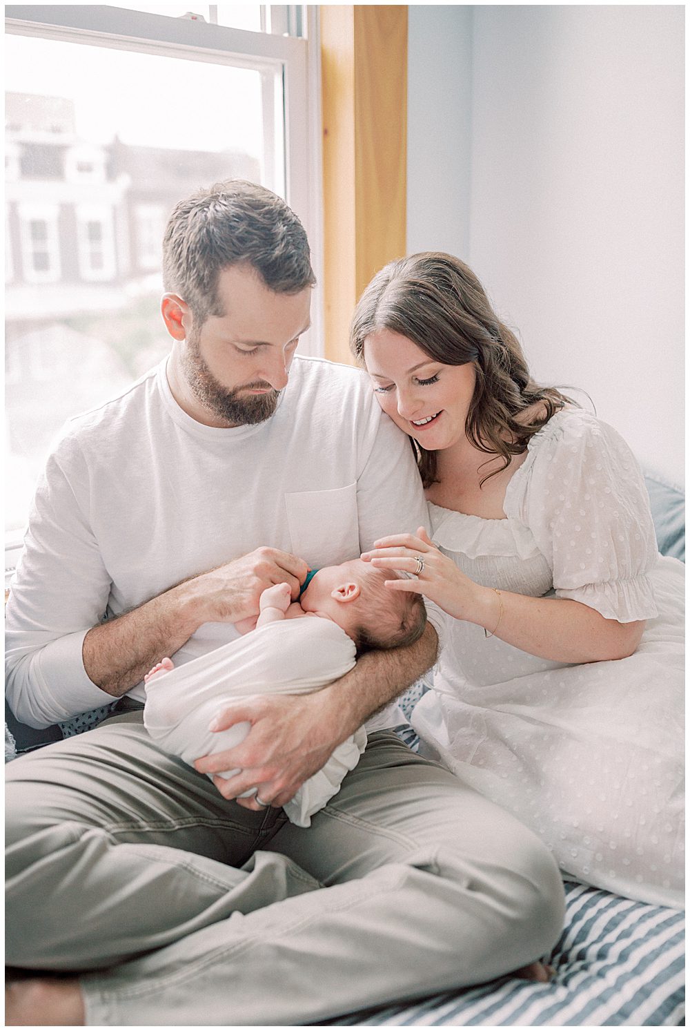 Dad sits on bed and holds his newborn son while wife leans against him, gently rubbing newborn's head during their Capital Hill newborn session.