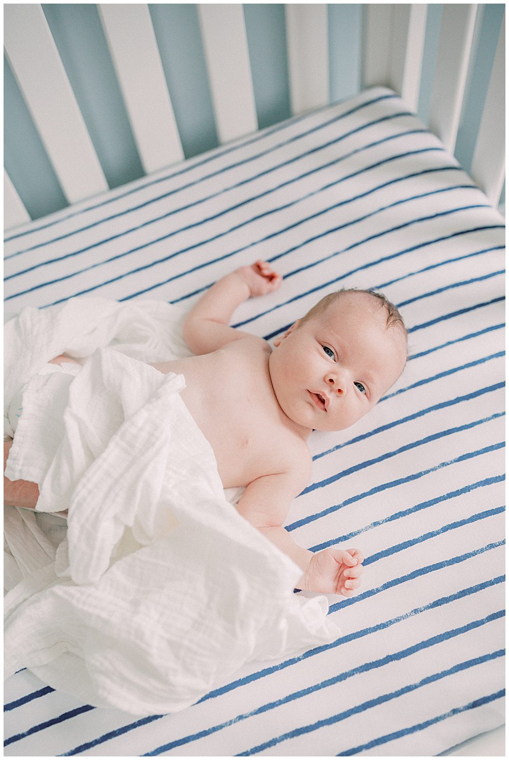 Baby boy loosely wrapped in a white swaddle lays in his navy and white striped crib