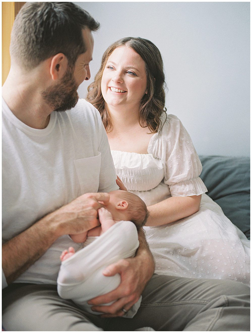 New mom smiles at her husband while he holds their new baby while they sit on their bed