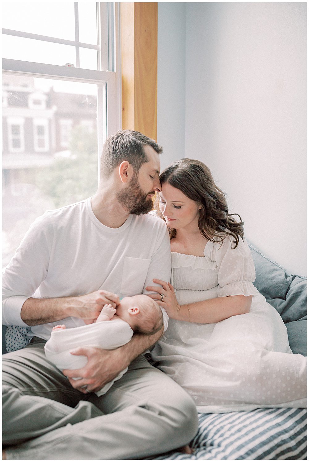 Father kisses the forehead of his wife while they sit on their bed holding their newborn baby during their Capital Hill Newborn Session