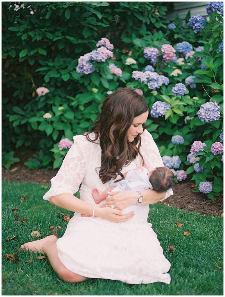 Brown-haired mother in white lace dress sits in front of a hydrangea bush holding her newborn baby.