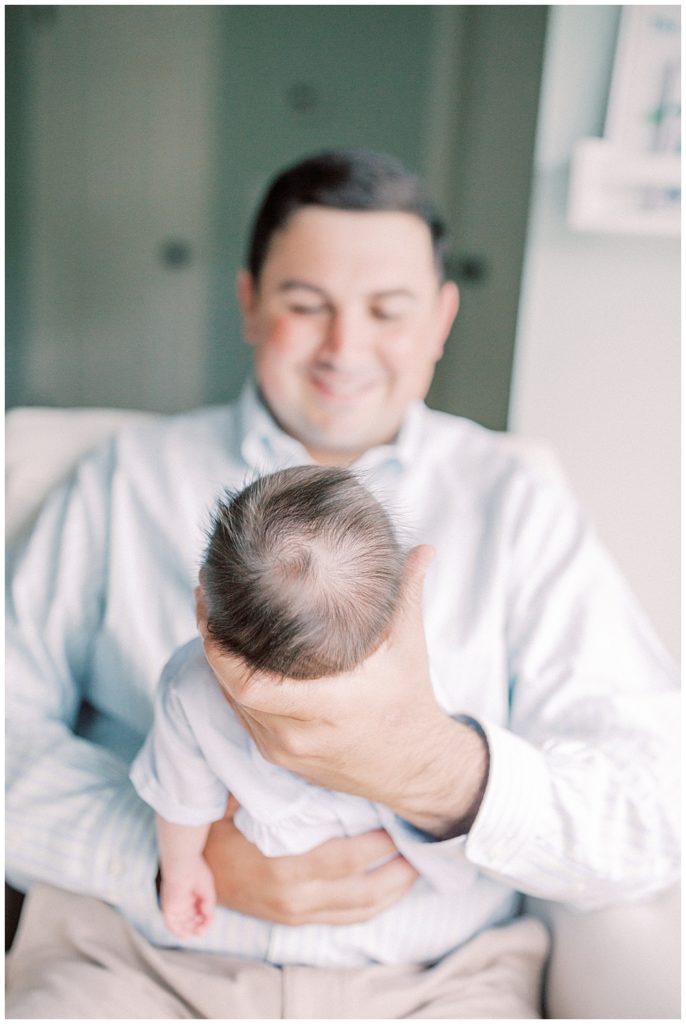 View. of fuzzy brown newborn hair on baby held by his father smiling down at him.