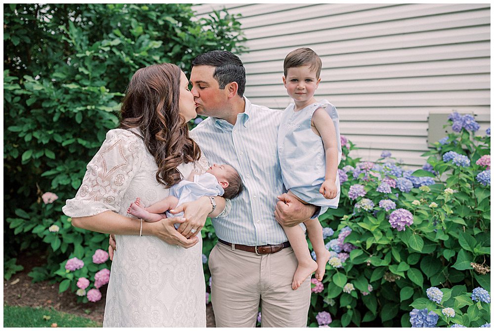 Mother and gather hold their toddler son and newborn and lean in for a kiss while standing in front of their hydrangea bush.