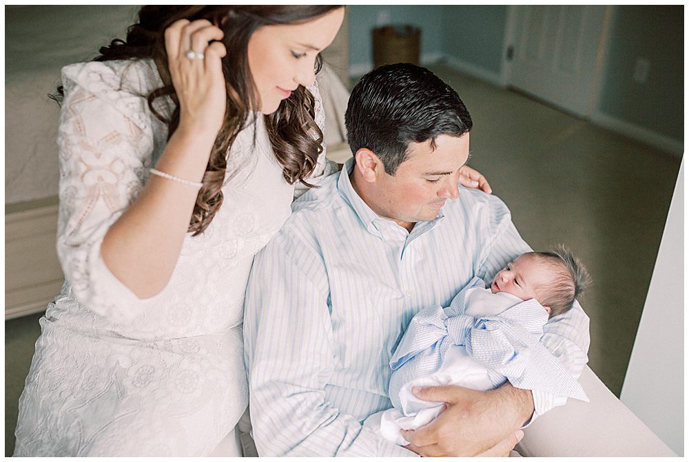 Mother and father sit together in chair holding and looking at their newborn baby boy. 