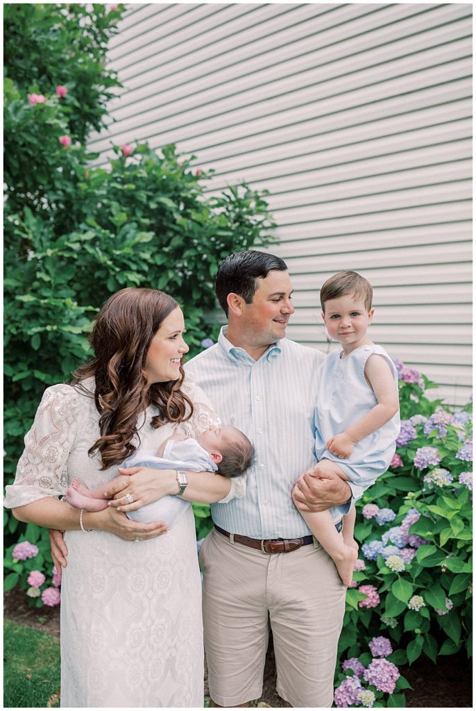 Mother and father smile at their toddler while holding both their toddler and newborn baby while standing in front of hydrangeas.