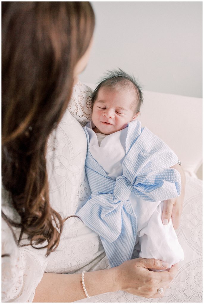 Newborn baby wrapped in light blue bow swaddle smiles while being held by mother during Fairfax newborn session.