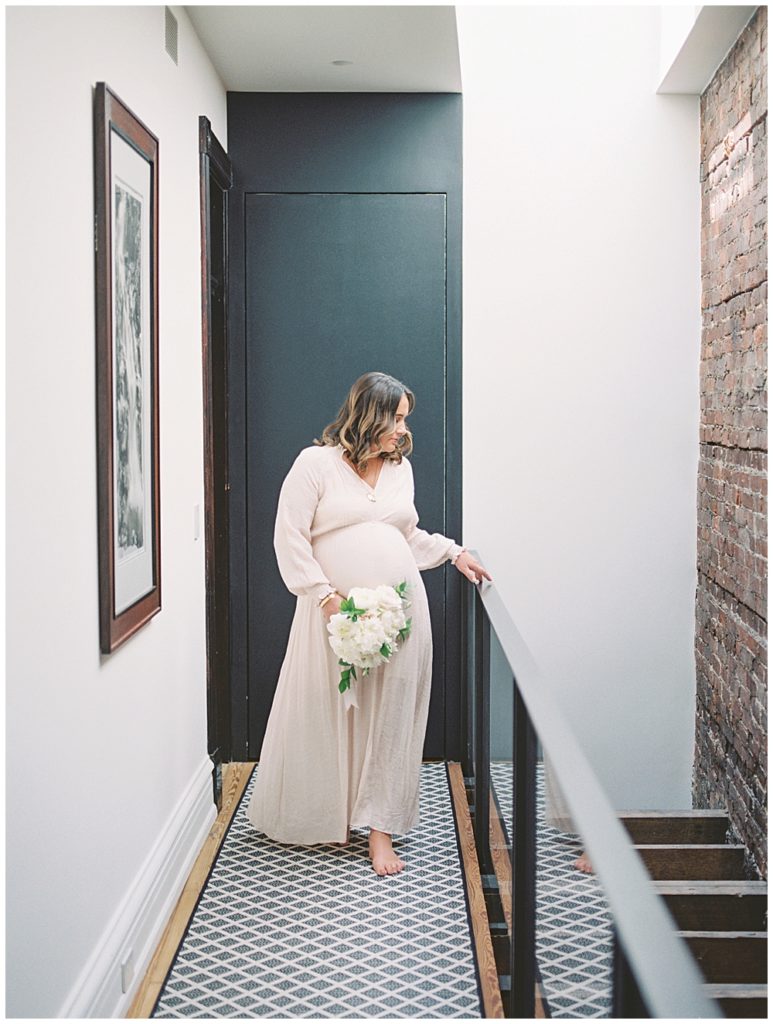 Pregnant mom walks through her hallway with one hand on her stair ledge and one holding a bouquet of white flowers during her in-home maternity session