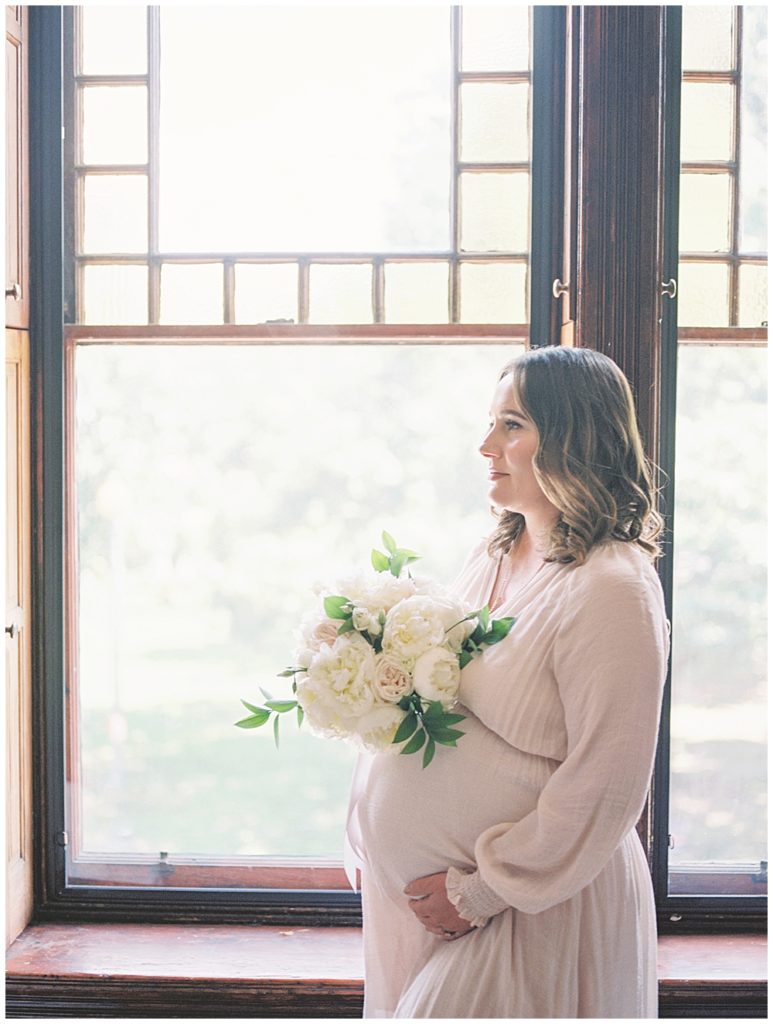 Pregnant mom stands in her window frame holding a bouquet of white roses with one hand below her belly