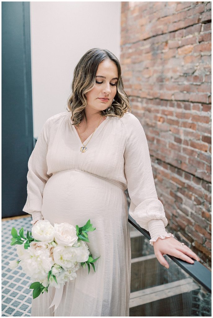 Expecting mother stands in a hallway holding a bouquet in one hand and placing her other hand on the guardrail