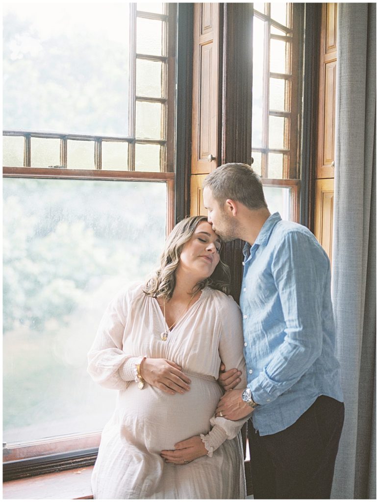 Pregnant mother sits in a large window while her husband kisses her head during their in-home maternity session