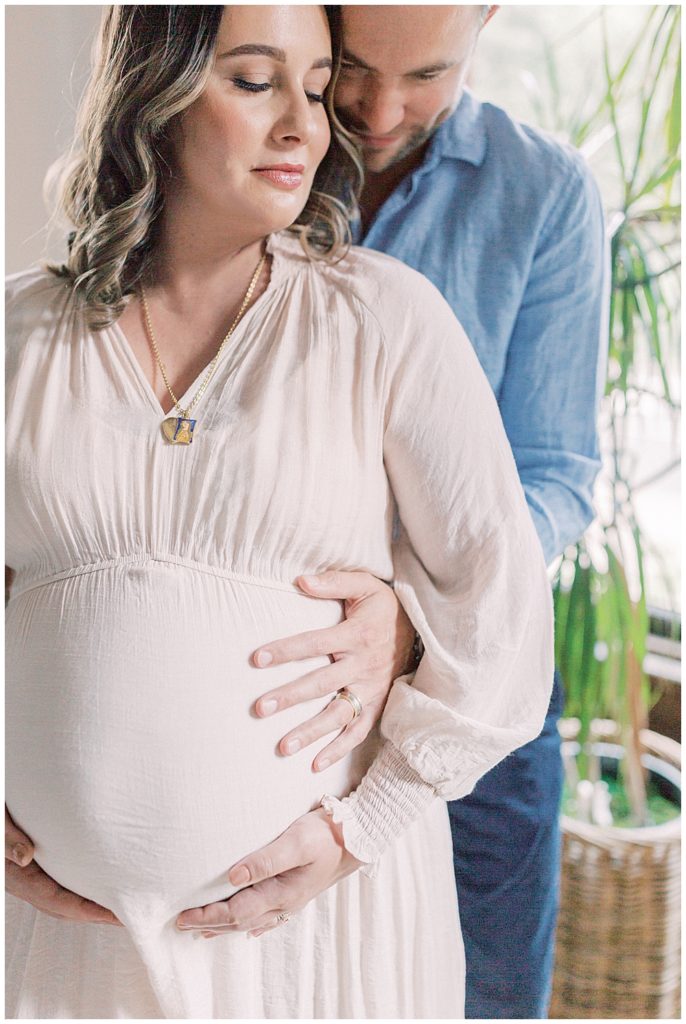 In-home maternity session where dad stands behind mom and holds mom's belly while they lean into each other
