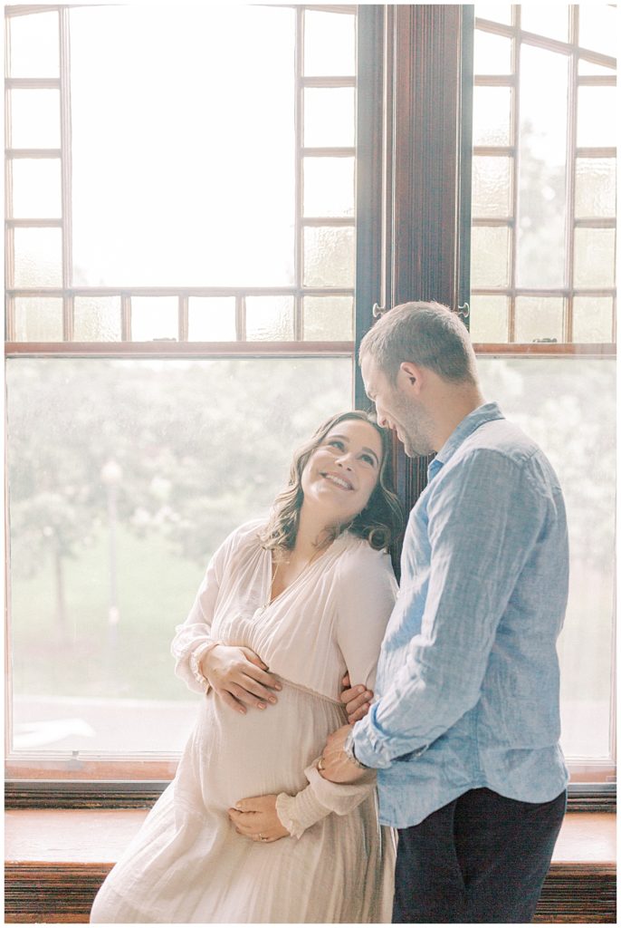 Pregnant wife sits in the windowsill while smiling up at her husband
