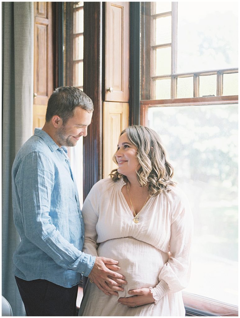 In-home maternity session in Washington, DC where pregnant mother sits in the window with her hand on her belly looking up at her husband