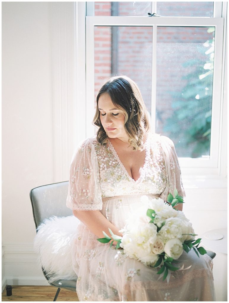 Pregnant mom sits in a chair in front of a bright window holding a bouquet and looking down off to the side