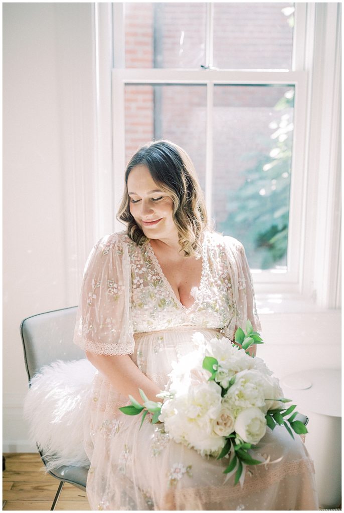 Pregnant woman in pink gown with white bouquet sits in front of a bright window and smiles