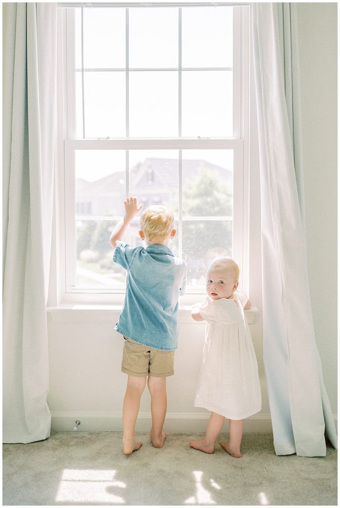Toddler girl stands with her bigger brother in the window and looks back at the camera.
