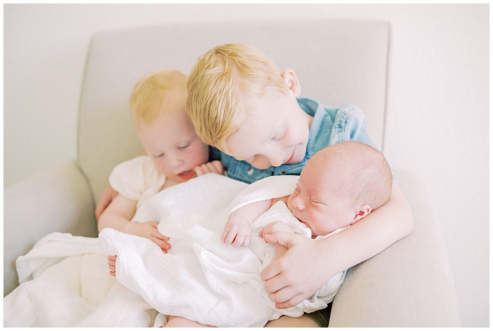 Red-haired toddler boy and girl hold and smile at their newborn baby brother.