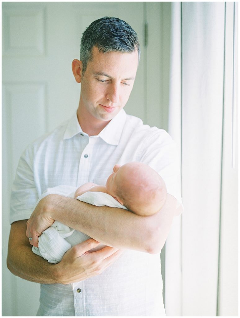 Father holds his newborn baby while standing ext to a window.